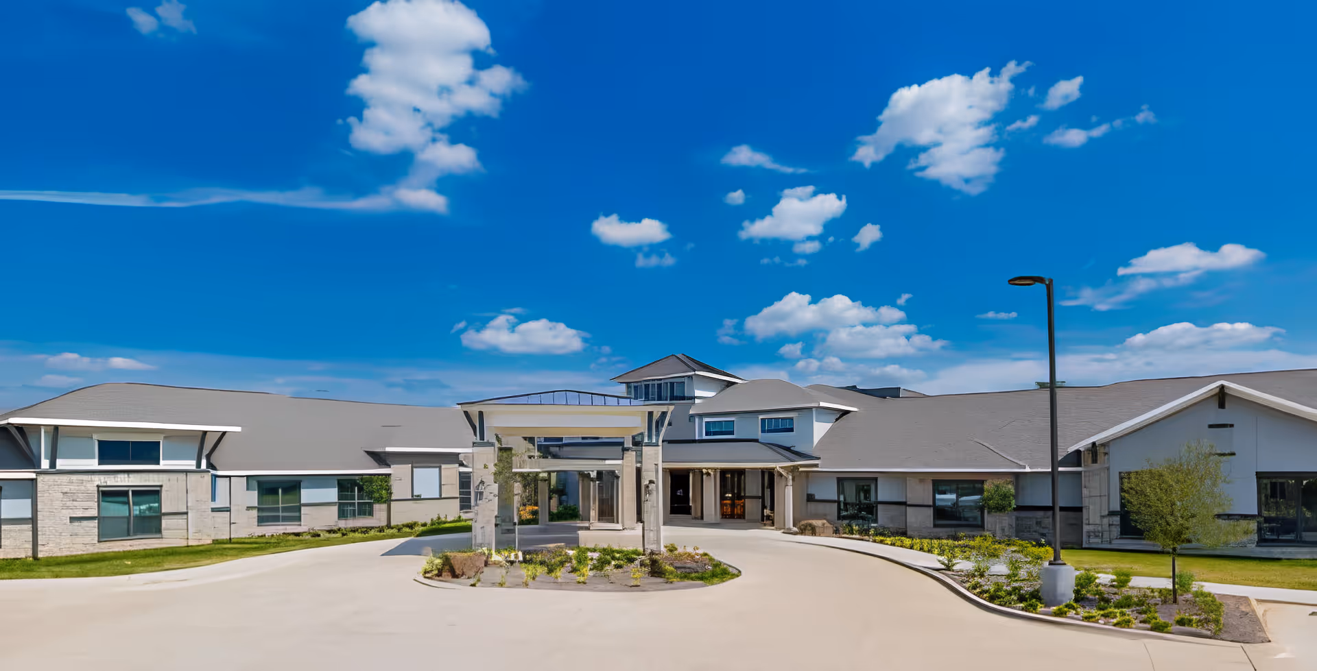 Front entrance of a low‑rise assisted living building with a circular driveway, landscaped island, and blue sky overhead.