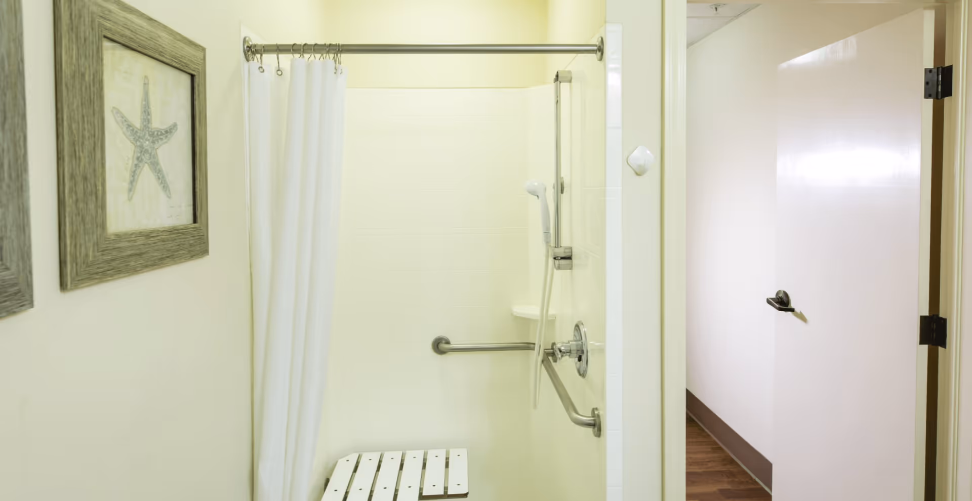 A bathroom shower area with a white shower curtain, a wall-mounted handheld showerhead, and a foldable shower seat. There are two framed pictures of starfish on the wall outside the shower. The bathroom door is open, showing a hallway with wooden flooring.