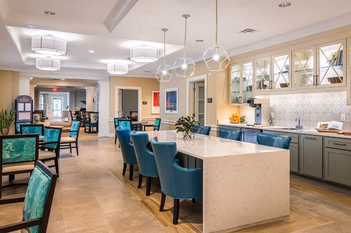 A bright and spacious dining area in a senior living facility featuring a long marble island with teal upholstered chairs, pendant lights hanging above, and a kitchen area with cabinets, a sink, and a coffee machine. Additional seating with green cushioned chairs and tables is visible in the background along with decorative artwork on the walls.