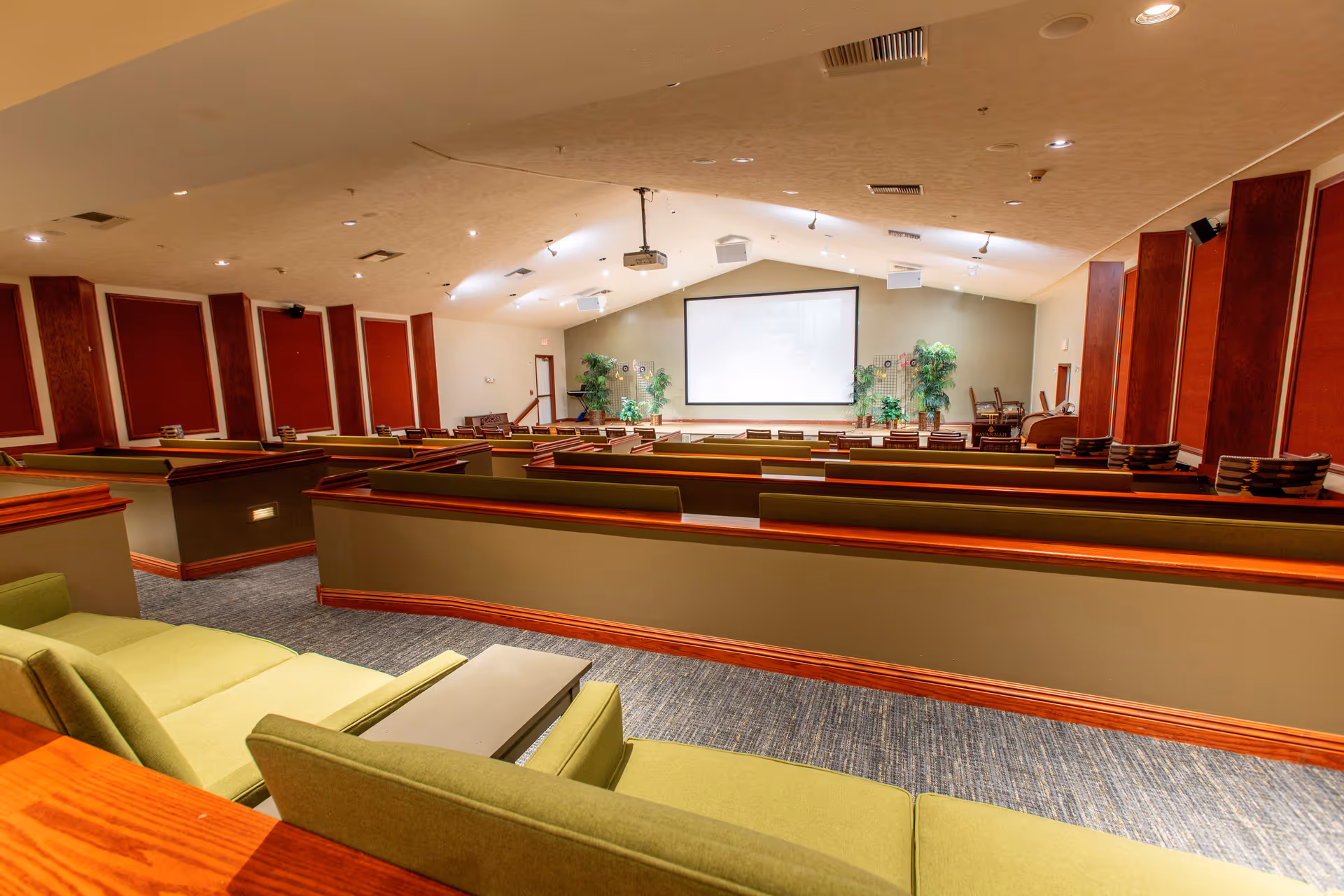 Interior view of a large auditorium-style room with tiered seating facing a projection screen.