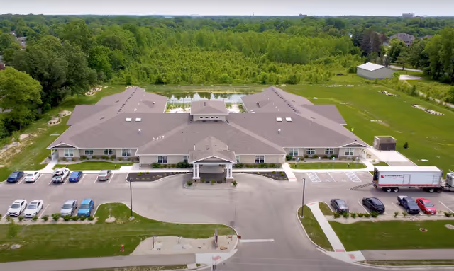 Aerial view of a single-story senior living facility named Lincolnshire Place of Decatur, surrounded by green trees and grass. The building has a large parking lot with several cars and a truck parked. The facility has a covered entrance and a pond behind it.