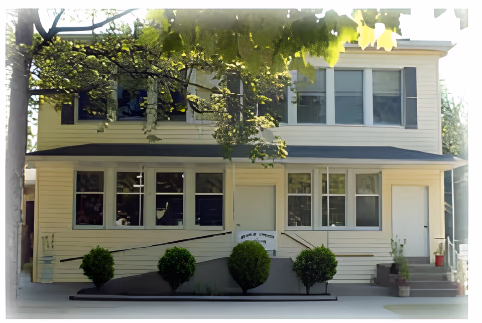 Front exterior view of a two-story yellow building with multiple windows, a black roof over the porch area, a ramp with handrails, and four small green bushes in front. There are stairs on the right side leading to a white door, and trees partially shading the building.