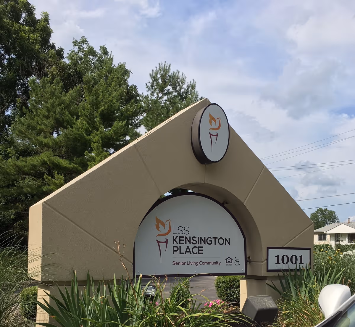 Outdoor sign for LSS Kensington Place Senior Living Community with the address number 1001, surrounded by greenery and trees under a partly cloudy sky.