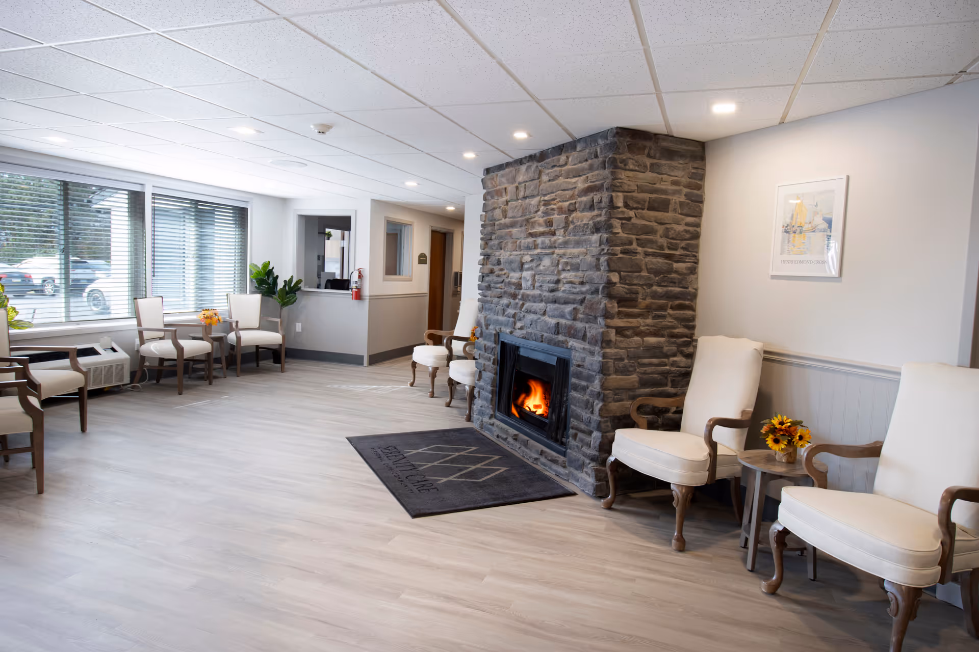 A bright and spacious common area with light wood flooring, a stone fireplace with a fire burning, several cushioned wooden armchairs arranged along the walls, a small table with a flower arrangement, large windows with blinds letting in natural light, and a reception window in the background.