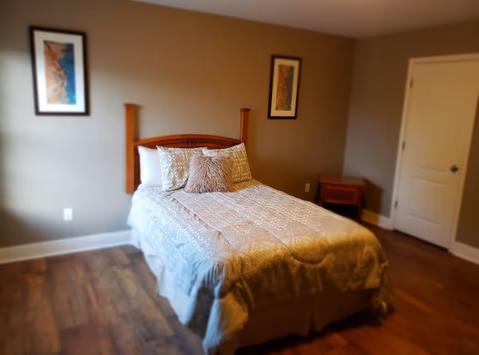 A bedroom with a neatly made bed featuring patterned bedding and three pillows, including a decorative furry one. The room has wooden flooring, beige walls, two framed pictures hanging above the bed, a wooden nightstand to the right, and a closed white door.