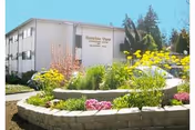 Exterior view of Sunrise View Retirement, Assisted Living, Rehabilitation & Skilled Nursing facility with a landscaped garden featuring colorful flowers and greenery in front of a two-story building under a clear sky.