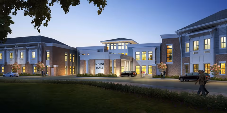 Front exterior of Indigo Hall assisted living building lit at dusk with entrance drive, parked cars, and a few people.