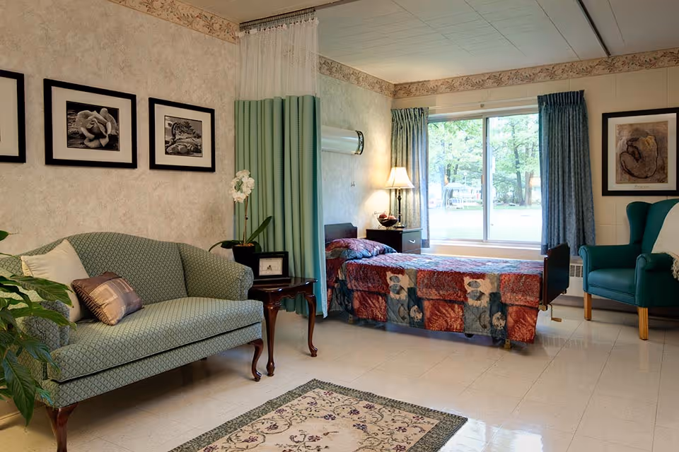 A cozy room with a single bed covered in a colorful quilt, a green upholstered loveseat with pillows, a green armchair, a wooden side table with a lamp, and a large window with curtains showing a view of trees outside. The walls are decorated with framed black and white artwork.