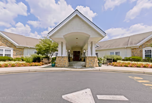 Covered porte-cochère entrance to a single-story senior living building with stone columns, benches, and landscaped beds under a partly cloudy sky.