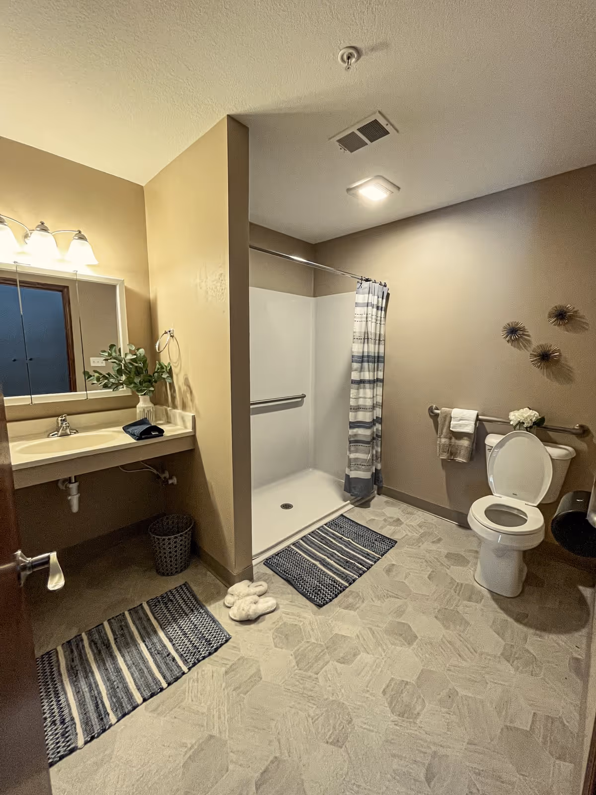 A clean and spacious bathroom with beige walls and hexagonal tile flooring. The room features a walk-in shower with a striped shower curtain, a white toilet with a towel hanging on a grab bar beside it, and a sink with a large mirror above it. There are decorative elements including a vase with greenery on the sink counter, wall art above the toilet, and two striped rugs on the floor along with a pair of white slippers.
