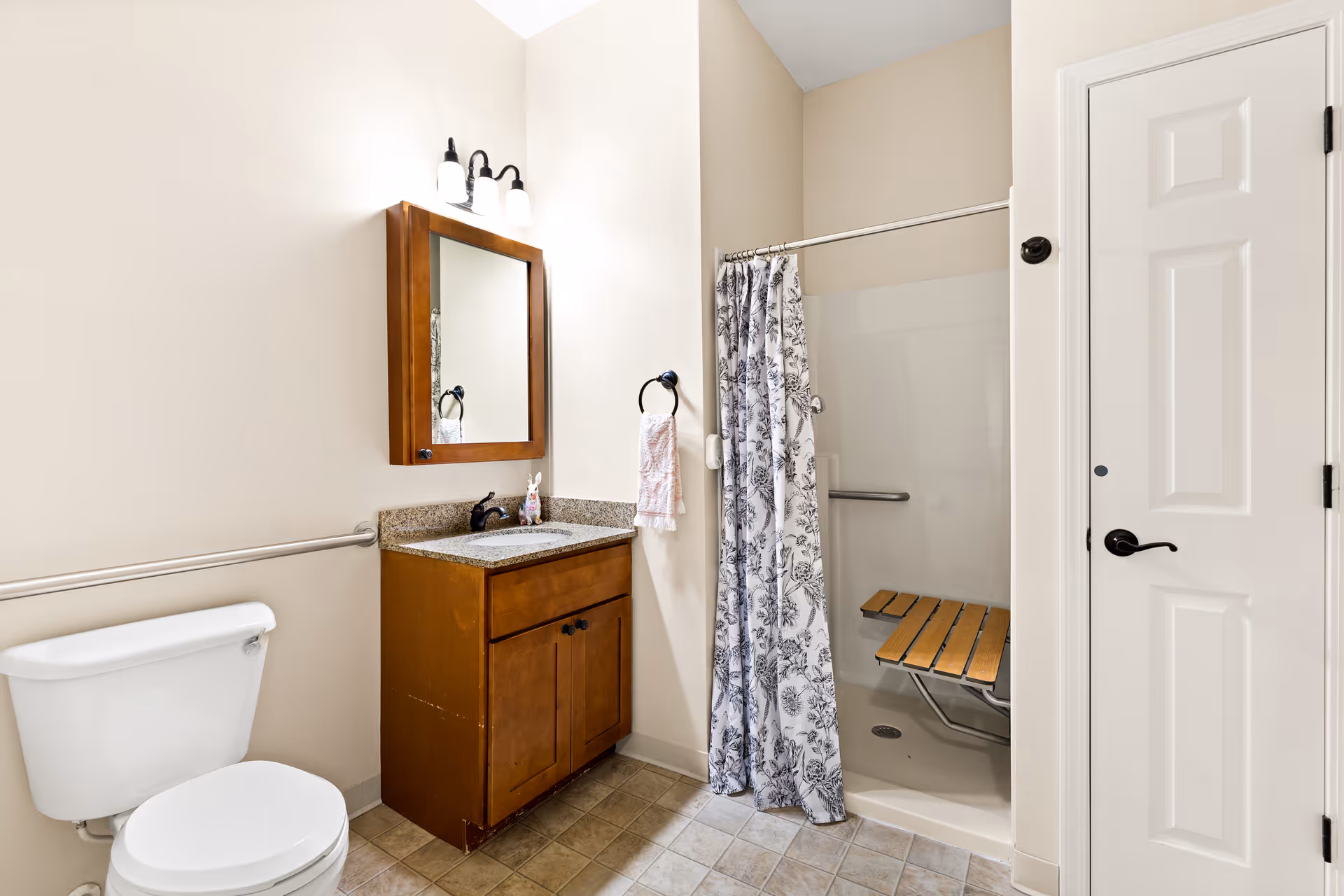 A bathroom with a white toilet, a wooden vanity with a granite countertop and a mirror above it, a towel ring with a hand towel, and a walk-in shower with a floral shower curtain and a wooden fold-down seat inside.