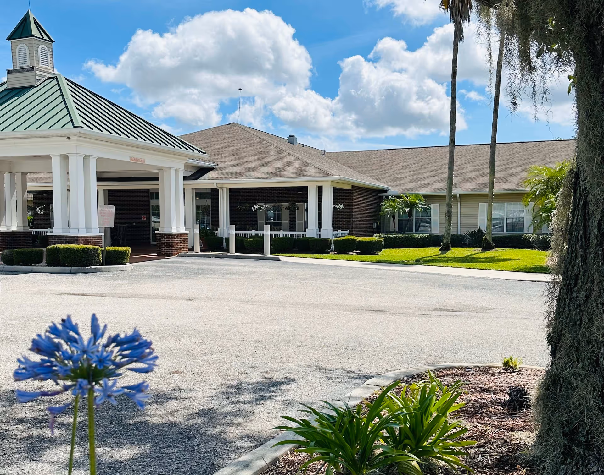 Front entrance of a single-story assisted living facility with a covered porte-cochere, landscaped grounds, and palm trees under a blue sky.