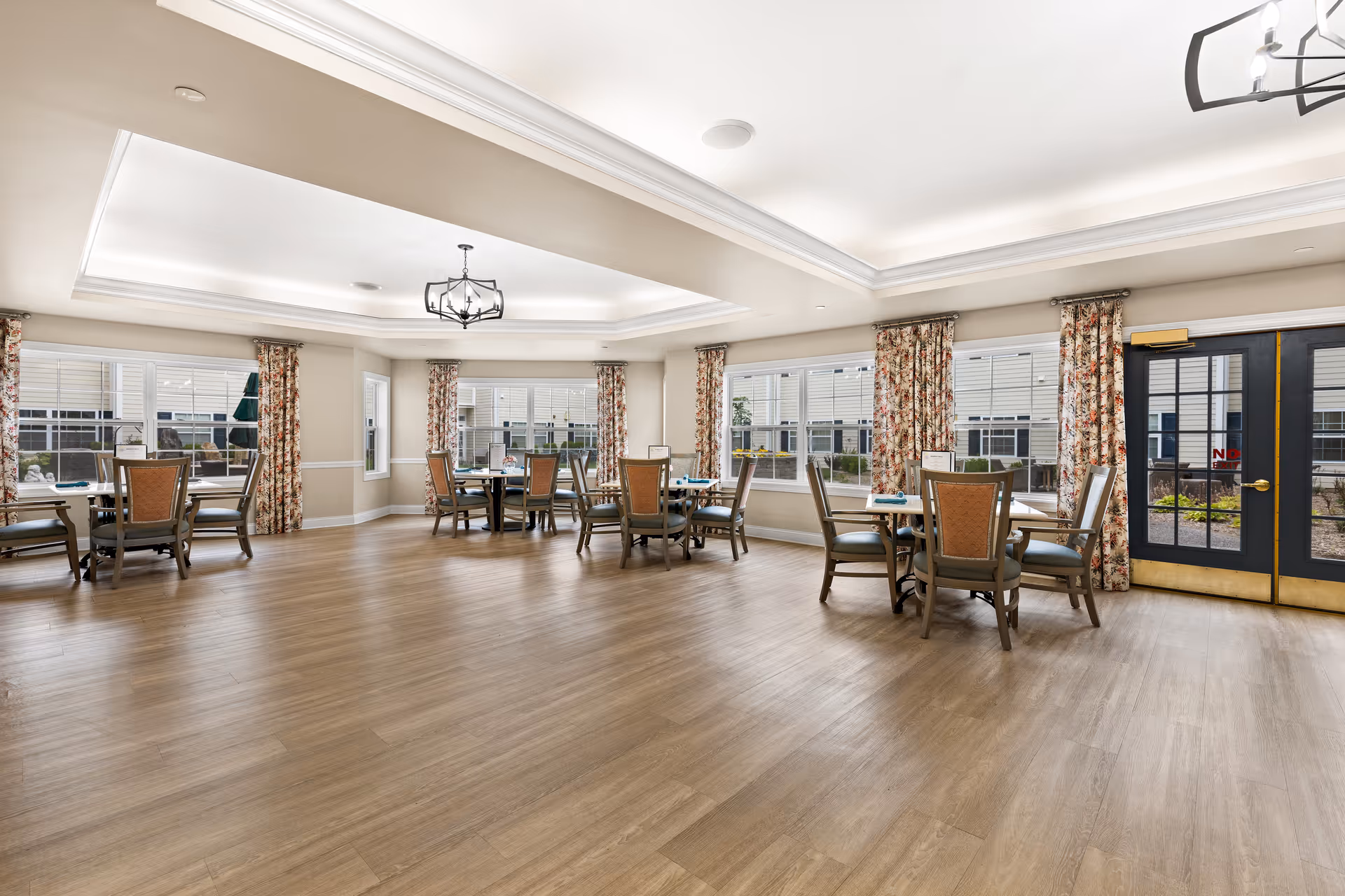 Spacious bright communal dining room with several round tables and chairs, large windows with floral curtains, and double doors.