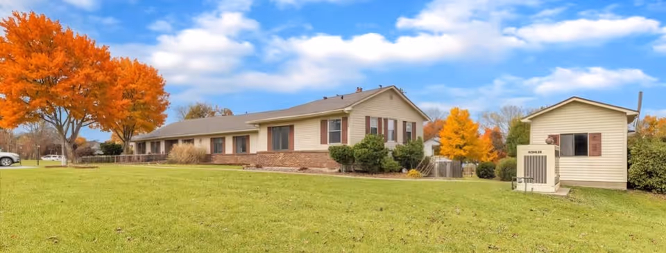 A single-story residential building with beige siding and brown shutters surrounded by green grass and trees with autumn-colored leaves under a partly cloudy blue sky.