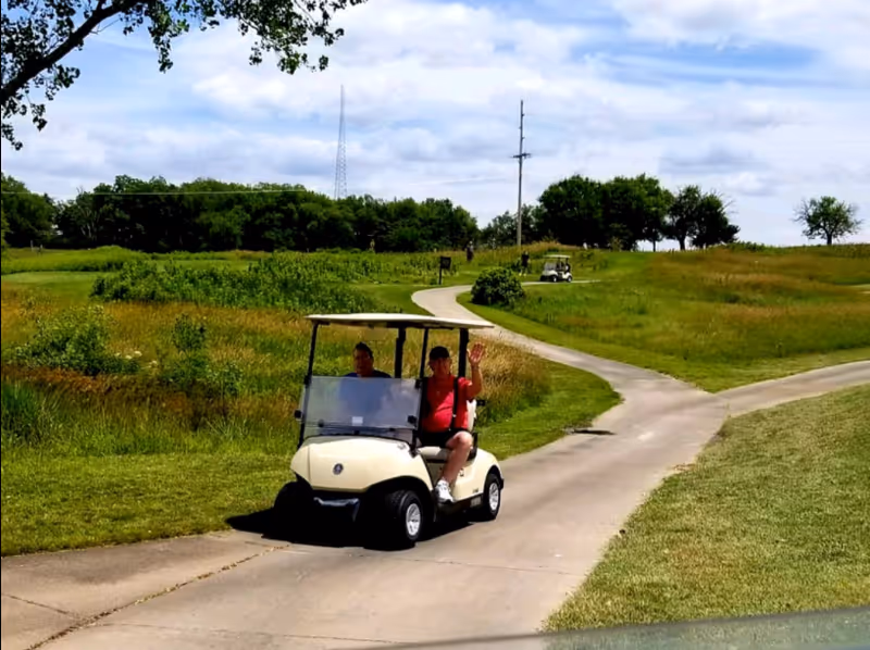 Two people ride a golf cart on a winding park path through open grassy fields.