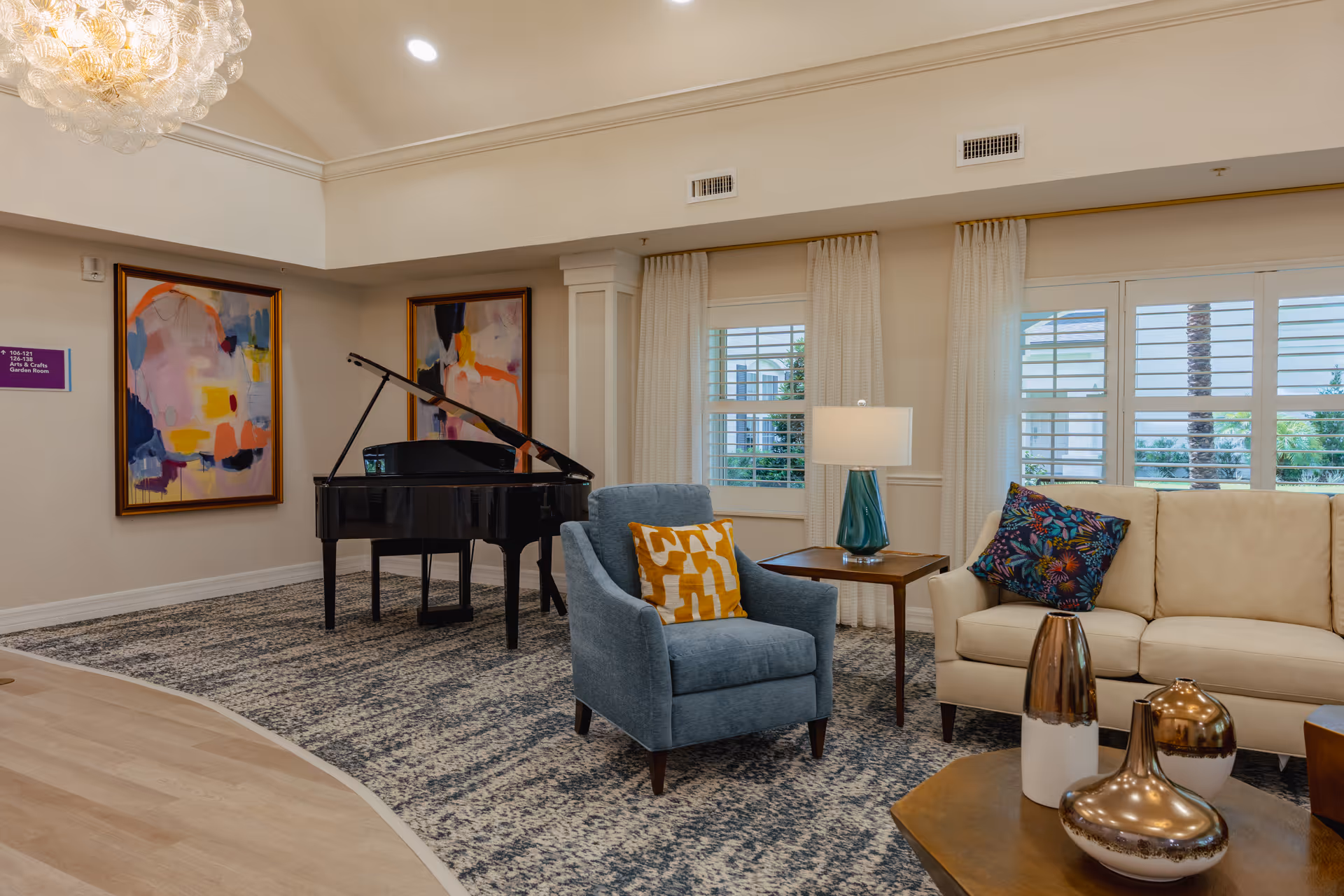 A cozy living room area in a senior living facility featuring a beige sofa with a colorful floral pillow, a blue armchair with a yellow patterned pillow, a wooden side table with a teal lamp, and a grand piano against the wall. The room has large windows with white curtains and two abstract paintings on the wall.