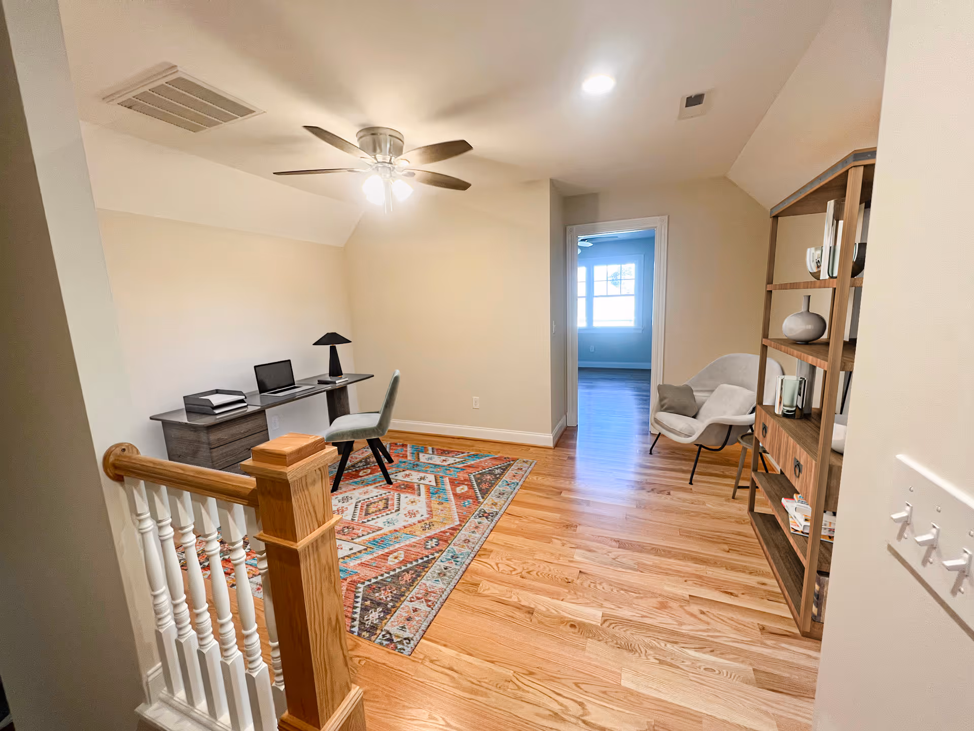 Bright upstairs landing used as a home office with a desk and chair, colorful rug, shelving unit, and an armchair by a doorway.