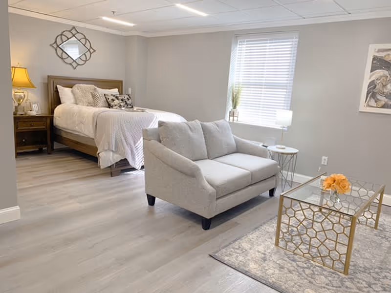 A bright and cozy senior living bedroom featuring a neatly made bed with white bedding and decorative pillows, a wooden nightstand with a lamp, a small gray loveseat, a glass coffee table with a gold geometric frame and a vase of orange flowers, a window with white blinds, and light gray walls and flooring.
