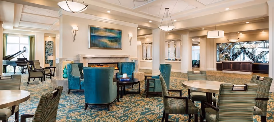 Bright senior living common area with round dining tables, striped chairs, teal armchairs around a fireplace and a grand piano in the background.