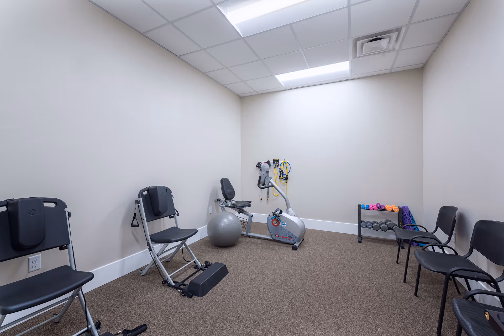 A small exercise room with beige walls and carpeted floor. The room contains two black exercise chairs, a recumbent exercise bike, a gray exercise ball, a rack with various dumbbells and yoga mats, and some resistance bands hanging on the wall. The ceiling has white tiles with fluorescent lighting.