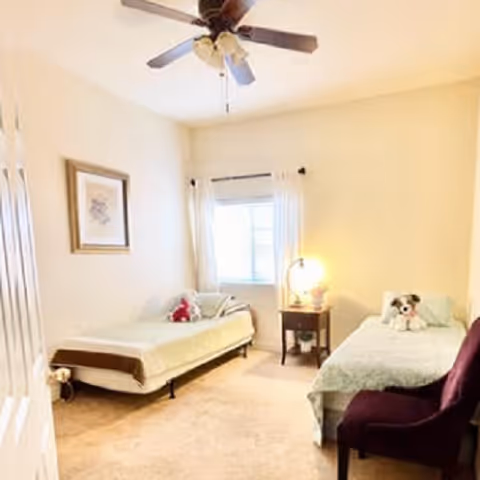 A bright bedroom with two single beds, each with a stuffed animal on top. There is a window with white curtains between the beds, a small wooden nightstand with a lamp, a ceiling fan, and a framed picture on the wall.