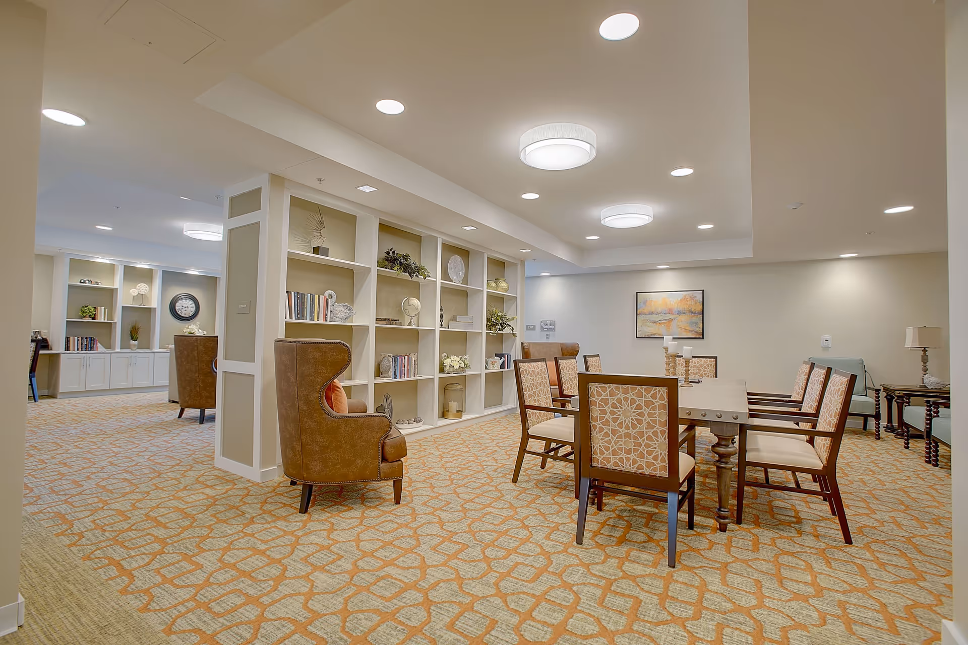 A spacious, well-lit common area with a large wooden table surrounded by six upholstered chairs with patterned backs. The room features a beige and orange patterned carpet, built-in white shelves filled with books and decorative items, and two brown leather wingback chairs. The walls are painted light beige, and there is a framed landscape painting on the far wall. Recessed ceiling lights and two round ceiling fixtures provide illumination.