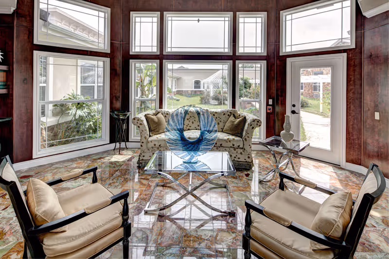 A bright and elegant living room with large windows and glass door letting in natural light. The room features a floral patterned sofa, two cushioned armchairs, a glass coffee table with a blue decorative sculpture, and a side table with a vase. The floor has a polished, multicolored marble-like finish and the walls are paneled in dark wood.
