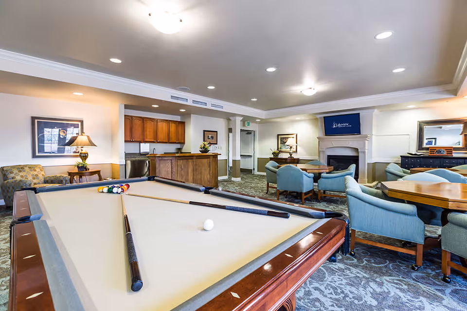 A spacious senior living common area featuring a pool table with cues and balls arranged, several round tables surrounded by blue upholstered chairs, a fireplace with a TV above displaying 'Lakeview Senior Living', wooden cabinetry, a lamp on a side table, and framed artwork on the walls.