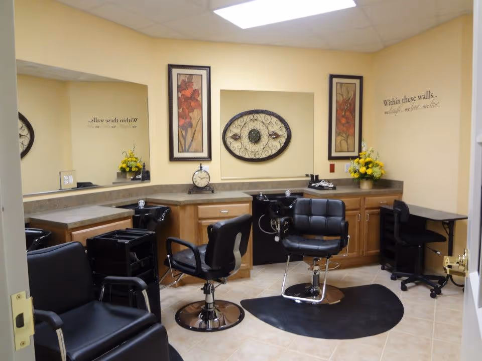 Interior view of a salon area with two black salon chairs in front of sinks and mirrors, a black salon trolley, wooden cabinets with countertops, decorative wall art, and a small desk with a chair. The walls are painted light yellow and there is a floral arrangement on the counter.