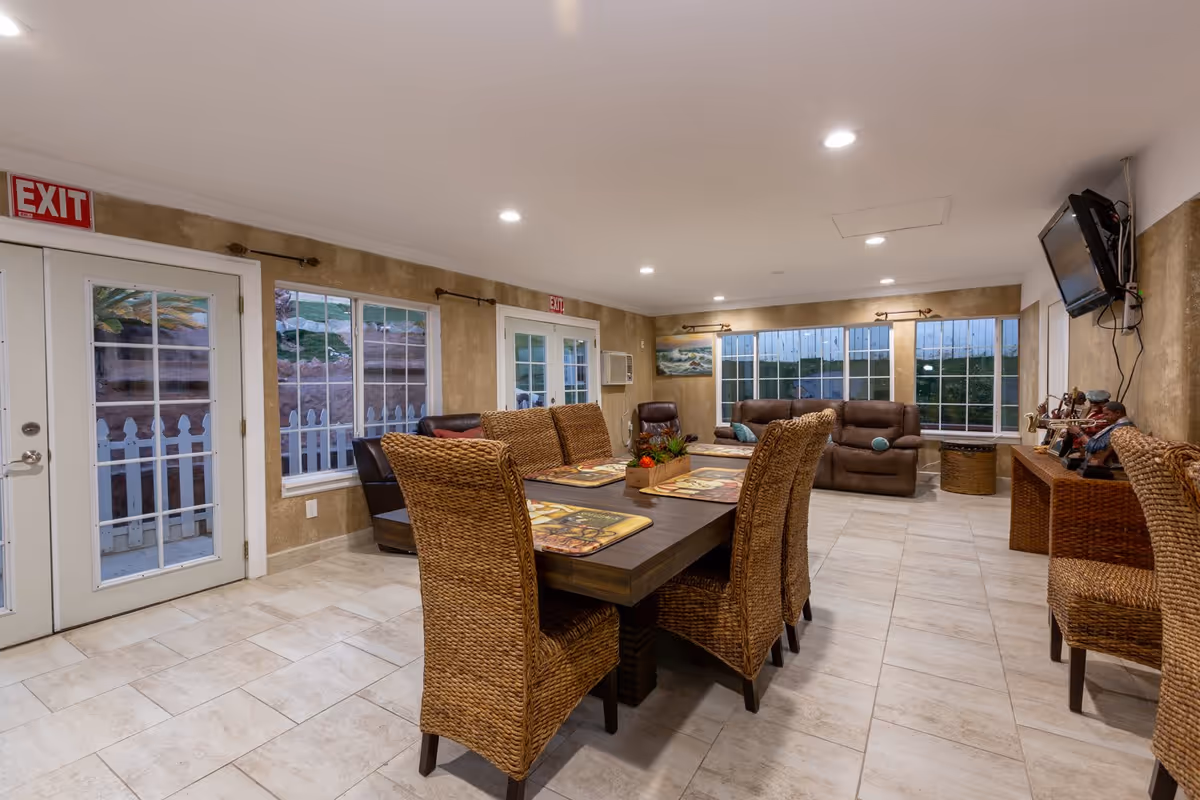 Spacious communal dining and lounge area with a long wooden table surrounded by wicker chairs, sofas, large windows and an exit door.