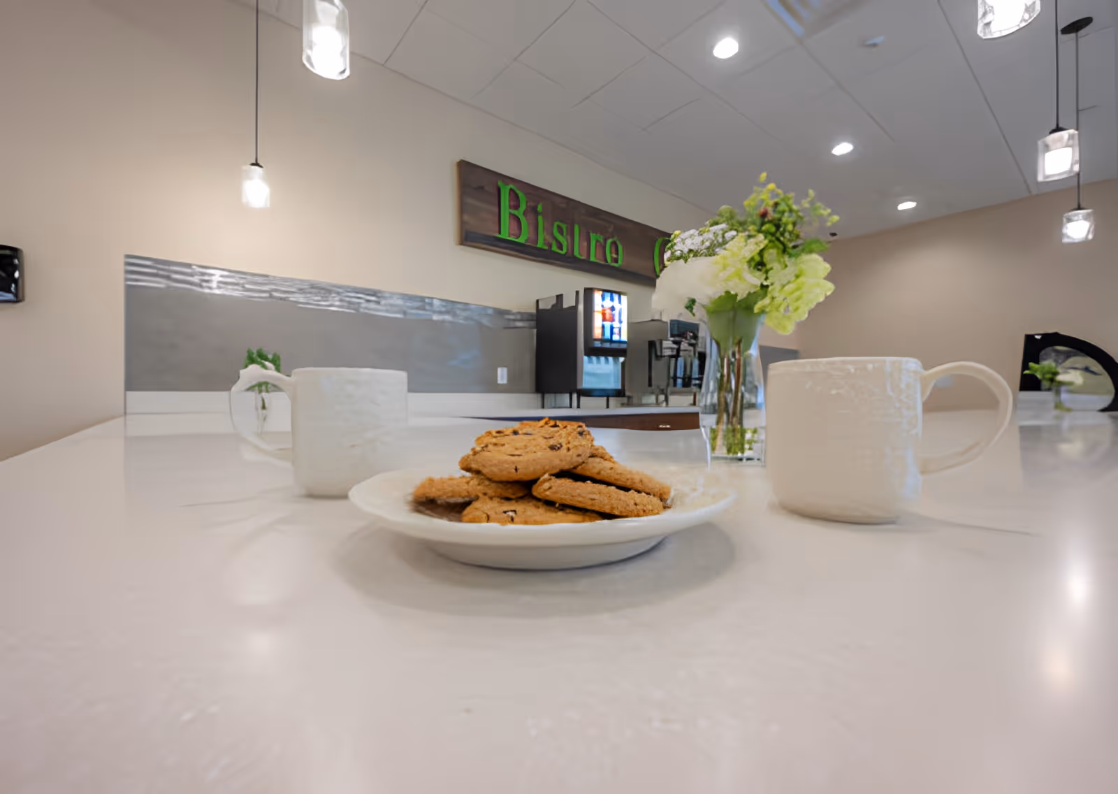 A plate of cookies and two mugs on a countertop in a bistro-style dining area with a drink machine and a vase of flowers in the background.
