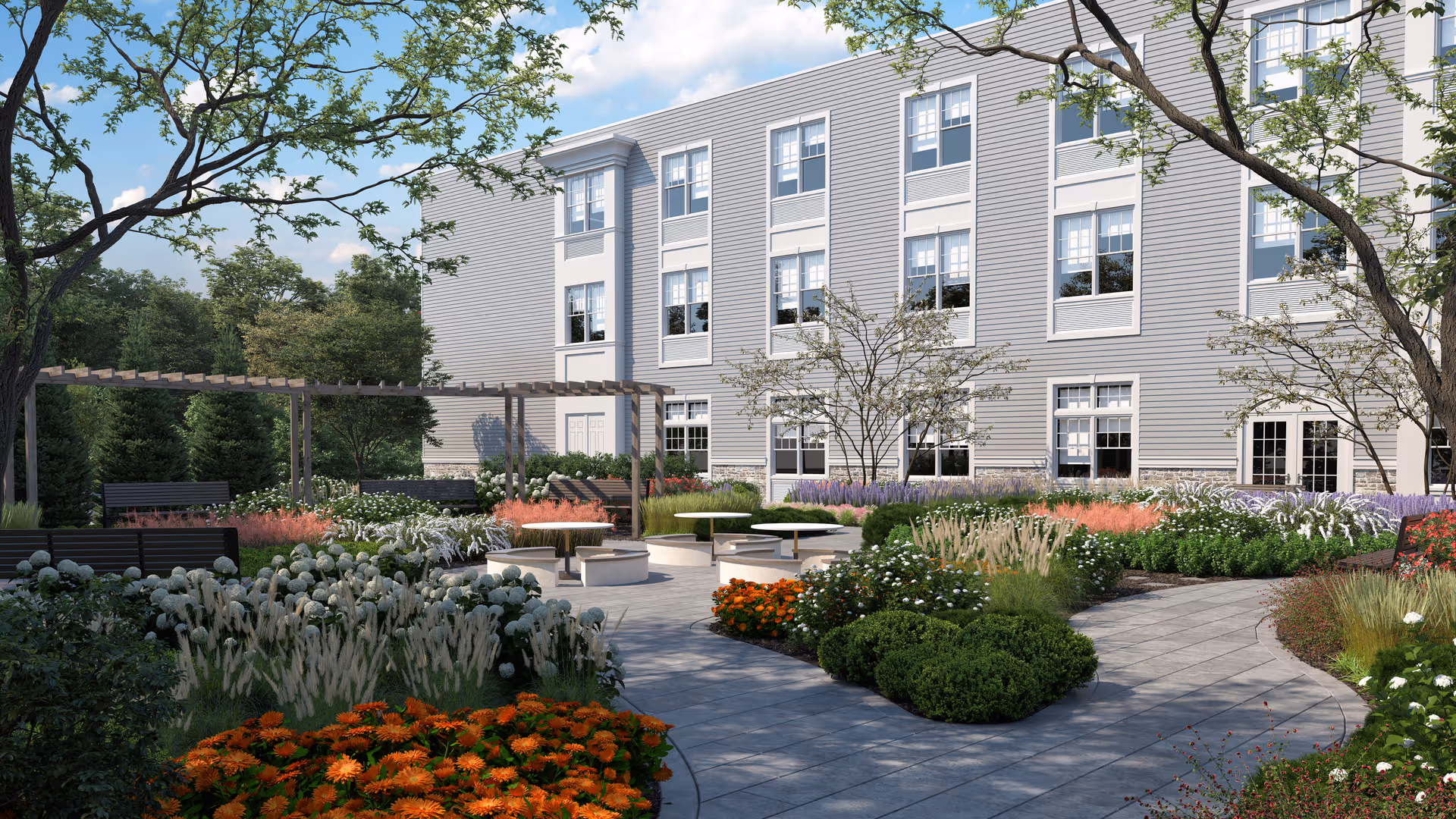 Outdoor garden area at The Bristal Assisted Living at Wayne featuring a paved walkway, various flowering plants and shrubs, benches, and a pergola structure with trees surrounding the space and a multi-story building in the background.
