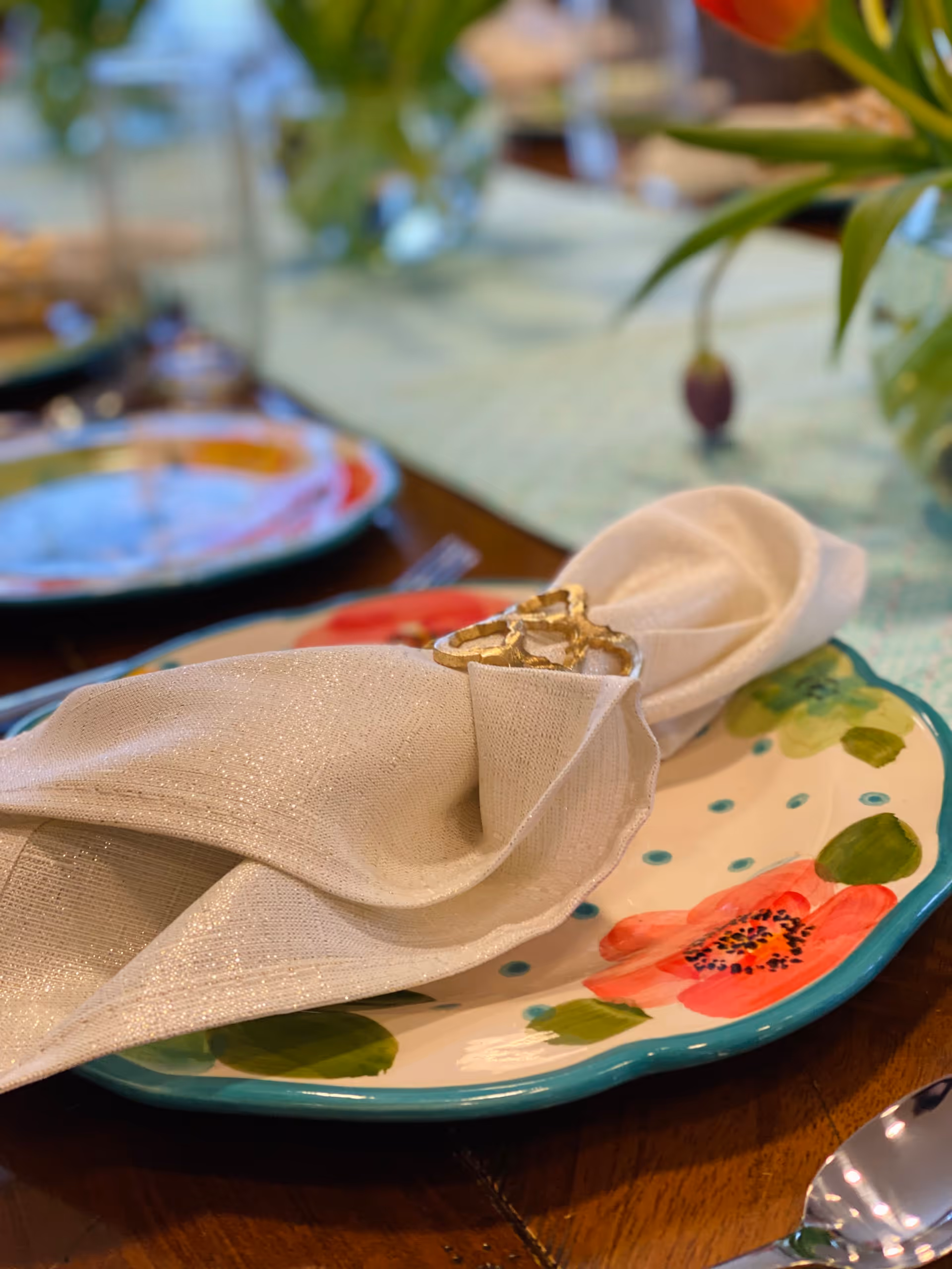 Close-up of a dining table setting featuring a white cloth napkin with a gold napkin ring placed on a colorful floral-patterned plate. The table has a green table runner and blurred background with other plates and greenery.