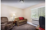 Living room with a recliner and loveseat (with a red pillow), side table and lamp, window with blinds and wall air conditioner, and a TV.