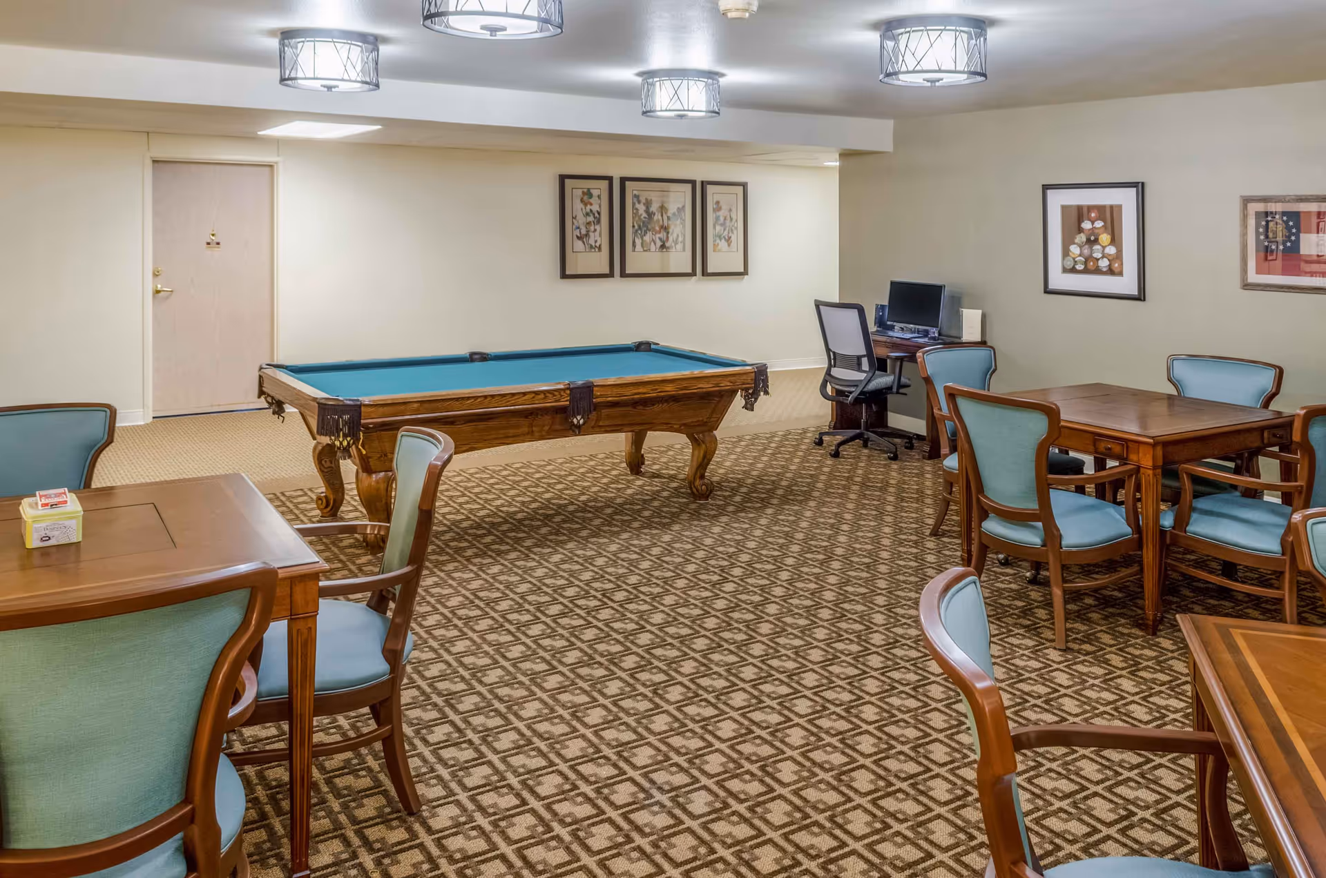 A recreational room with a pool table in the center, surrounded by several wooden tables and chairs with light blue upholstery. There is a computer workstation with a chair against the far wall, and framed artwork hanging on the walls. The room has patterned carpet flooring and ceiling lights.