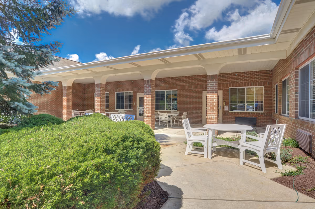 Covered brick patio courtyard with white tables and chairs, columns, windows, and shrubs under a blue sky.