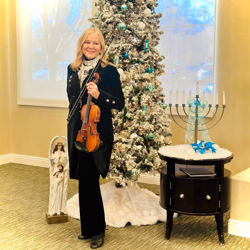 A woman holding a violin and bow stands indoors next to a decorated Christmas tree with white and teal ornaments. To the right is a menorah with white candles and a blue ribbon on a small round table. To the left of the woman is a statue of two angels. The room has beige walls and a window with a white shade.