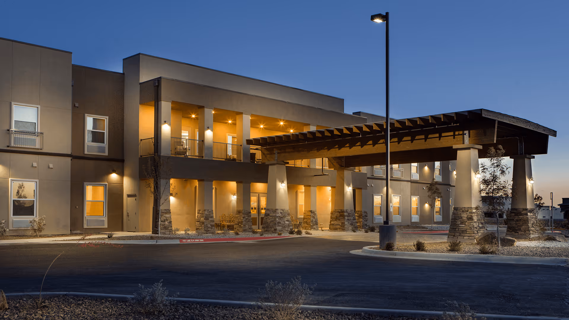 Exterior view of The Legacy at Santa Fe senior living facility at dusk, showing a two-story building with illuminated windows and a covered entrance supported by stone pillars. The sky is clear and a streetlamp is lit near the entrance.