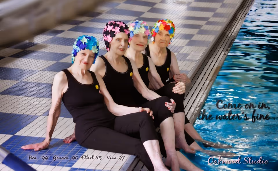 Four elderly women wearing black swimsuits and colorful swim caps sitting by the edge of an indoor swimming pool with their feet in the water. The pool area has a tiled floor with a blue and beige checkerboard pattern.