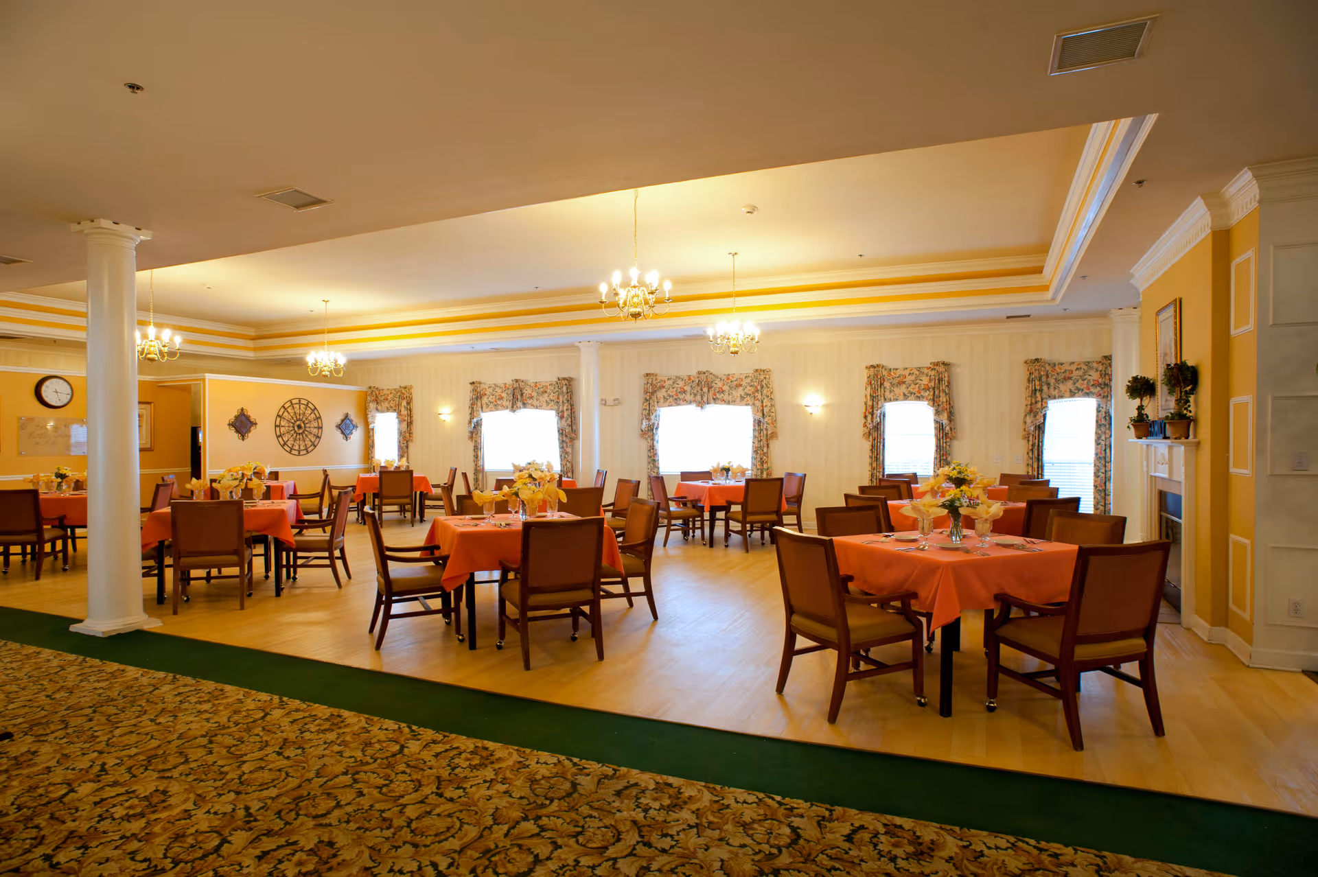 Spacious dining room with round tables covered in coral tablecloths, chairs, chandeliers, and floral centerpieces.