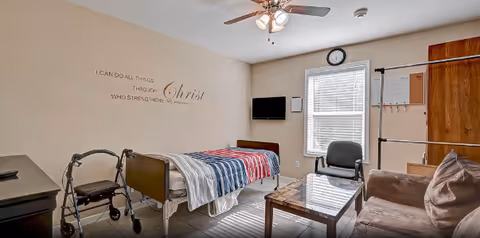 A well-lit assisted living bedroom with a single bed covered in a striped blanket, a walker beside the bed, a small TV mounted on the wall, a chair, a sofa, a coffee table, a clock above the window, and a religious wall decal that reads 'I CAN DO ALL THINGS THROUGH Christ WHO STRENGTHENS ME. Philippians 4:13'.