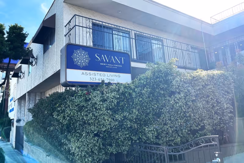 Exterior view of the Savant of West Hollywood assisted living facility building with a blue and white sign displaying the facility name and phone number, surrounded by greenery and a metal fence.