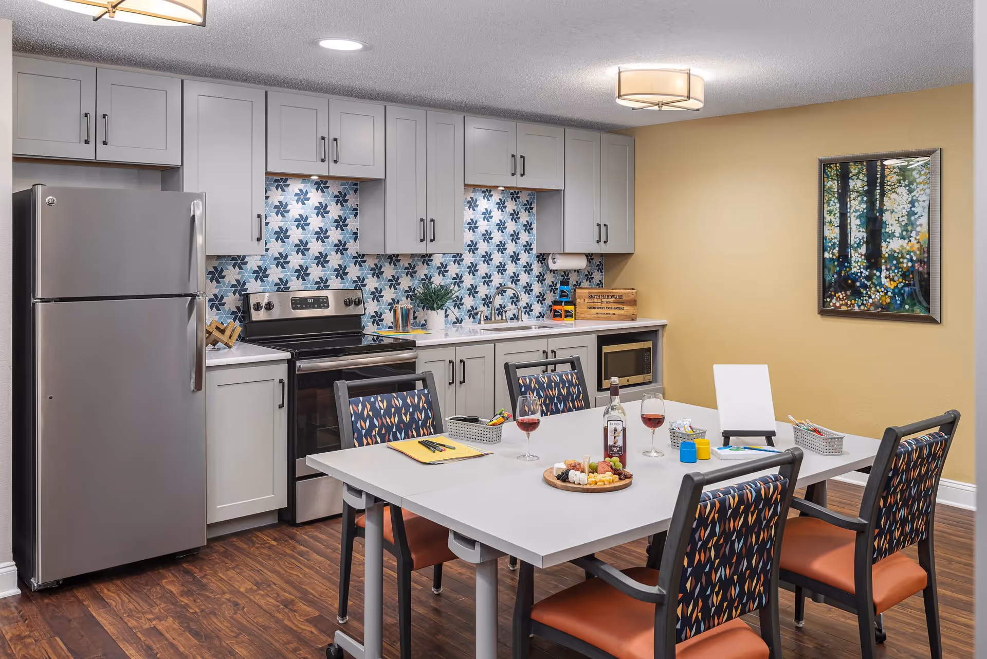 Communal kitchen and dining area with stainless-steel appliances, a patterned tile backsplash, and a table set with chairs and snacks.