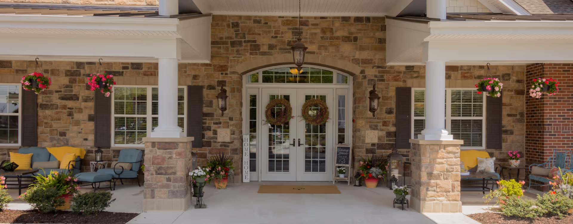 Front entrance of a senior living facility with stone and brick exterior walls, white double doors adorned with wreaths, two large white columns, hanging flower baskets, outdoor seating with cushions, and various potted plants and decorations.
