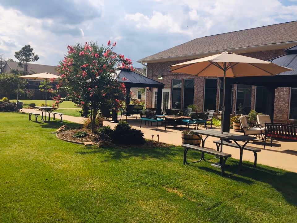 Outdoor patio area at a senior living facility with green grass, flowering tree, picnic tables with umbrellas, benches, and cushioned seating near a brick building under a partly cloudy sky.