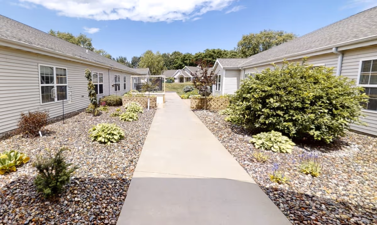 A paved walkway between two single-story buildings with beige siding, surrounded by landscaped areas with rocks, small bushes, and plants under a partly cloudy sky.