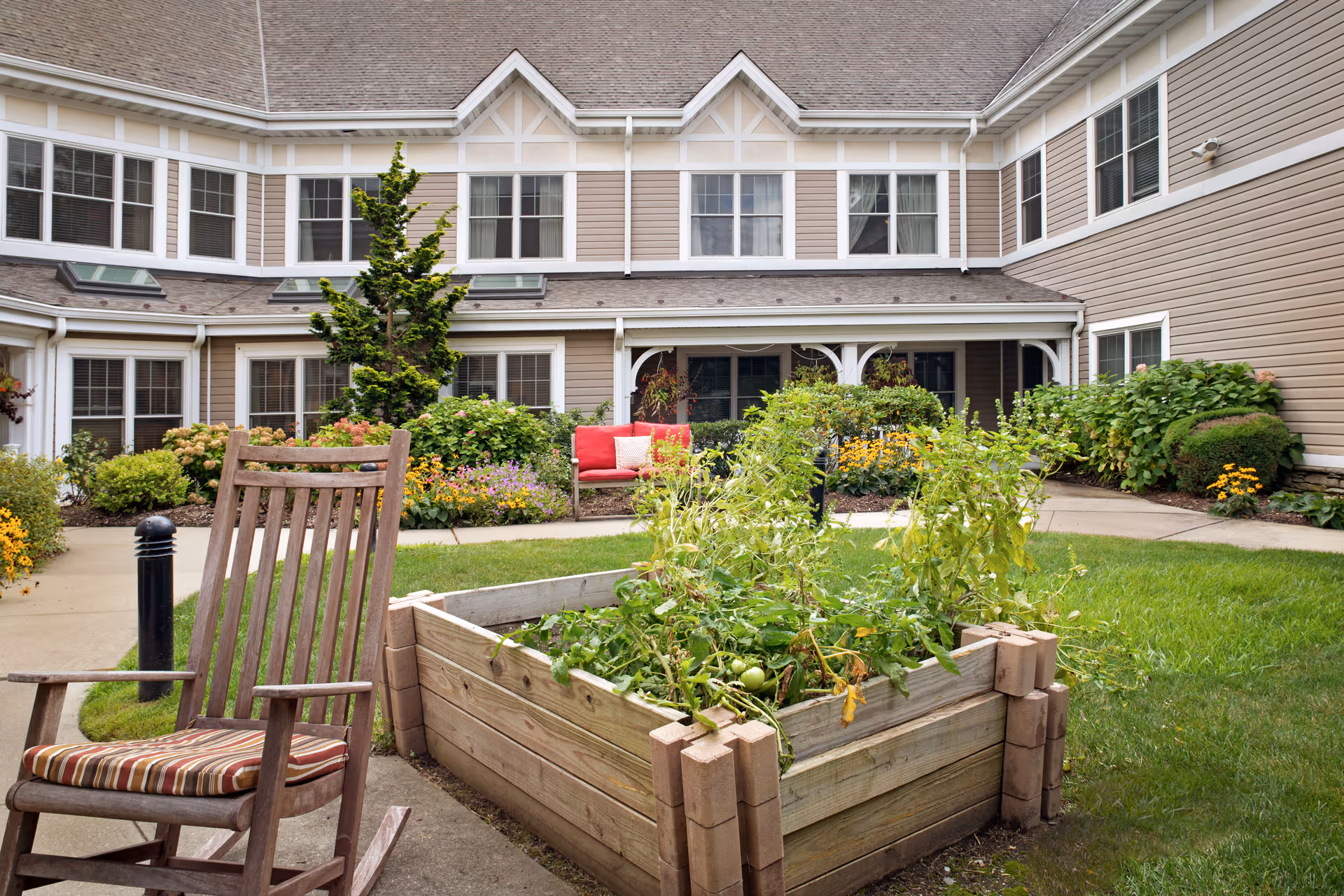 Courtyard garden with a raised wooden planter, a striped-cushion rocking chair, and red outdoor seating in front of a two-story senior living building.