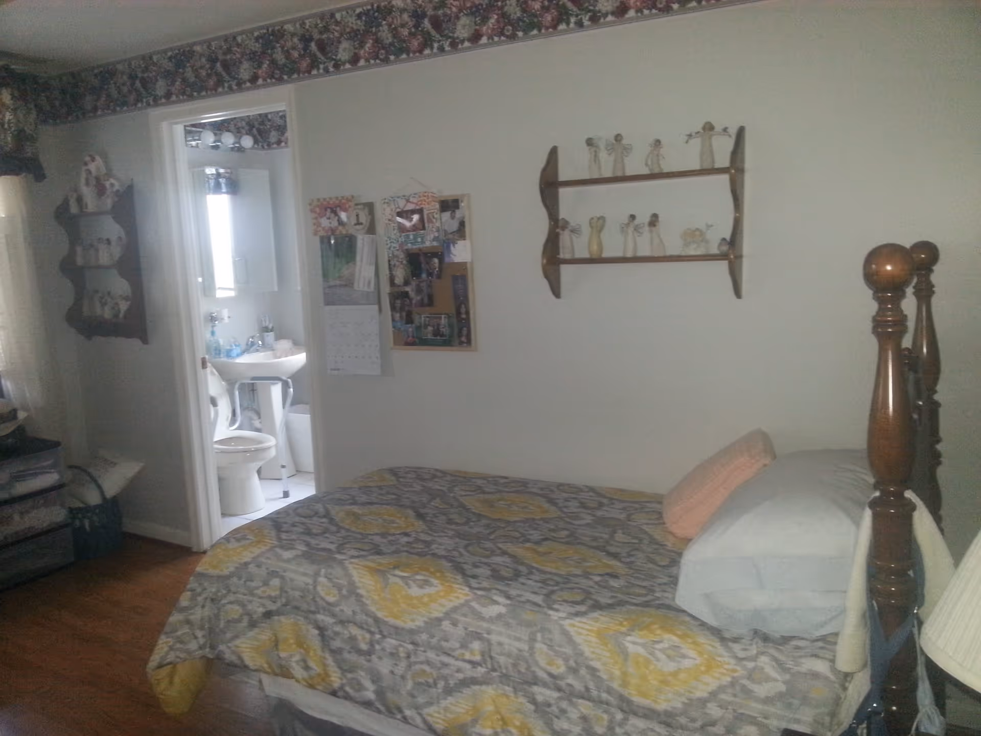 A bedroom with a wooden bed frame and a bed covered with a patterned quilt in shades of gray and yellow. There are pillows stacked at the head of the bed. On the wall above the bed is a wooden shelf displaying several small angel figurines. To the left, there is a doorway leading to a bathroom where a toilet, sink, and mirror are visible. The bedroom walls are decorated with a floral border near the ceiling and a bulletin board with photos and a calendar. A small wooden shelf with decorative items is mounted on the wall near the bathroom door.