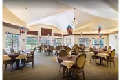 A spacious dining room with multiple round and rectangular tables covered with beige tablecloths. Each table is surrounded by cushioned chairs with wooden frames. The room has large windows allowing natural light to fill the space, and decorative hanging light fixtures from the high ceiling. The walls are painted in light colors, creating a warm and inviting atmosphere.