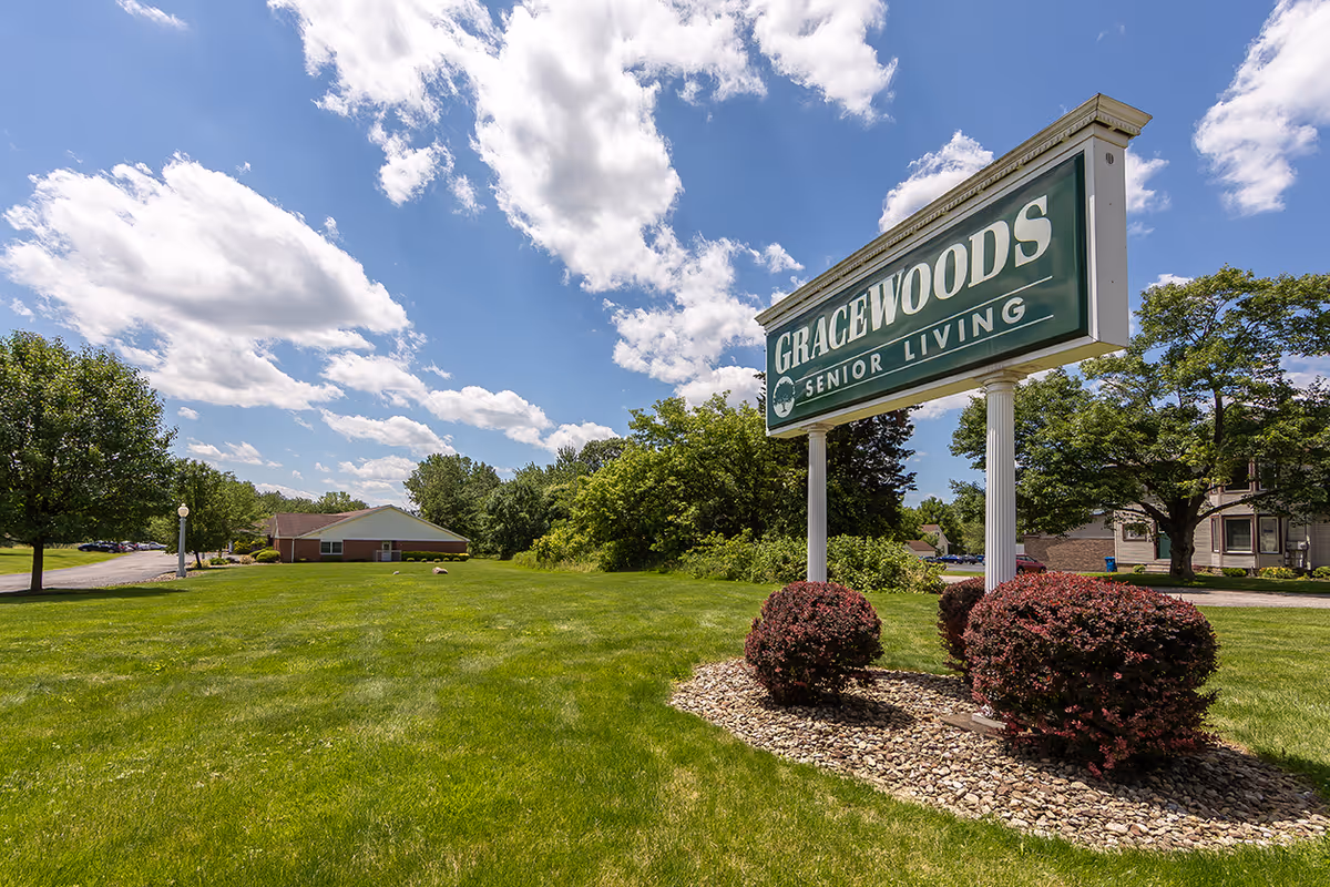 A large green lawn with neatly trimmed bushes and trees under a partly cloudy blue sky. A prominent sign reads 'GRACEWOODS SENIOR LIVING' in white letters on a green background. In the distance, there is a single-story brick building and a driveway with parked cars.